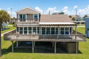 Two-story beach house on stilts with a large wraparound wooden deck and stairs under a blue sky.
