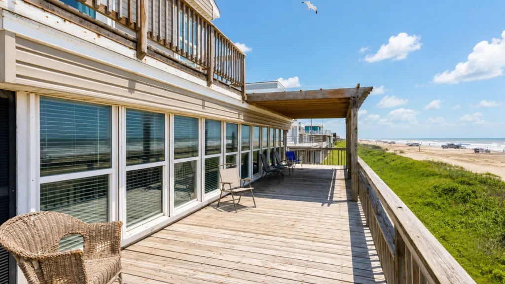 Wooden beachfront deck with wicker chair and glass windows, offering a view of the sandy beach and blue ocean under a clear sky.