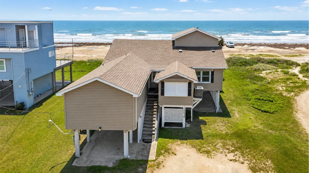 Aerial view of beige beachfront houses on stilts with a sandy beach and blue ocean beyond to the horizon.