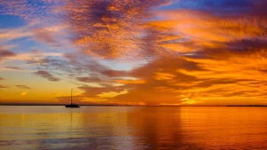 Colorful sunset over calm water with orange, gold, and blue clouds; a small sailboat silhouette near the left-center on the horizon.