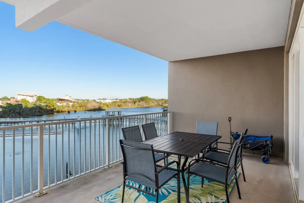 Balcony dining area with a black metal table and chairs, beige wall, and a railing overlooking a river and blue sky.