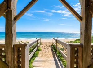 Wooden boardwalk leads to a sunny beach and blue ocean under a clear sky.
