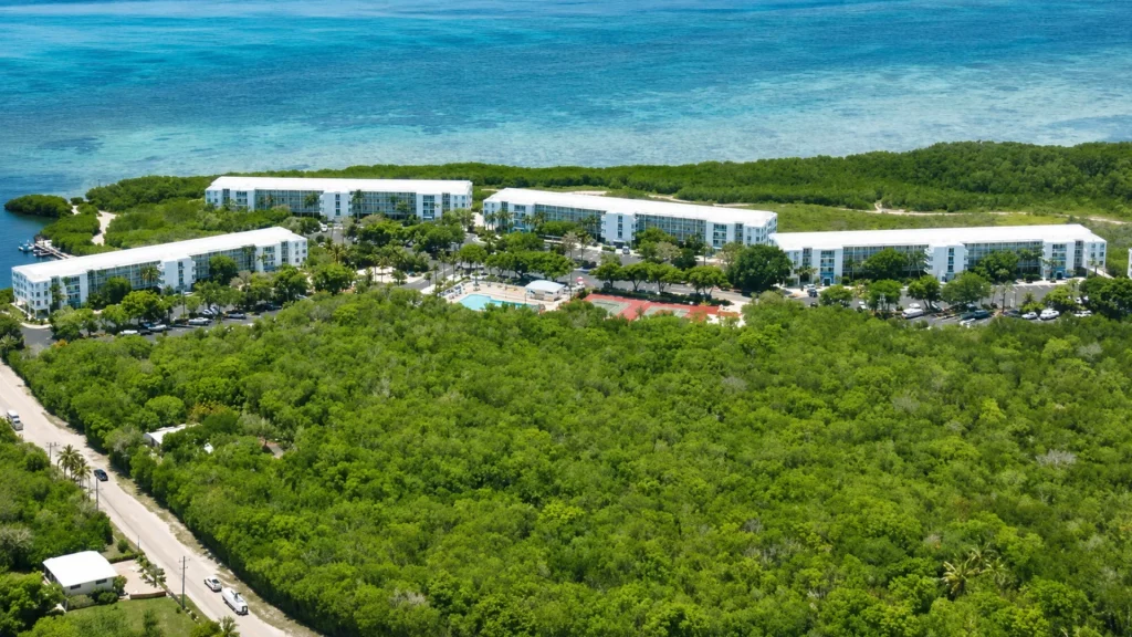 Aerial view of a white seaside resort with multiple low-rise buildings amid dense green mangroves, a central pool and tennis courts, and a turquoise ocean in the background.