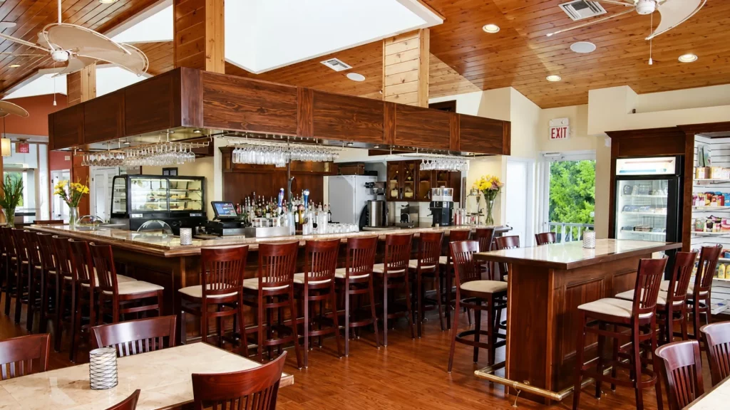 Interior of a bright bar with a long wooden counter, rows of stools, and glassware hanging overhead beneath a wood-paneled ceiling.