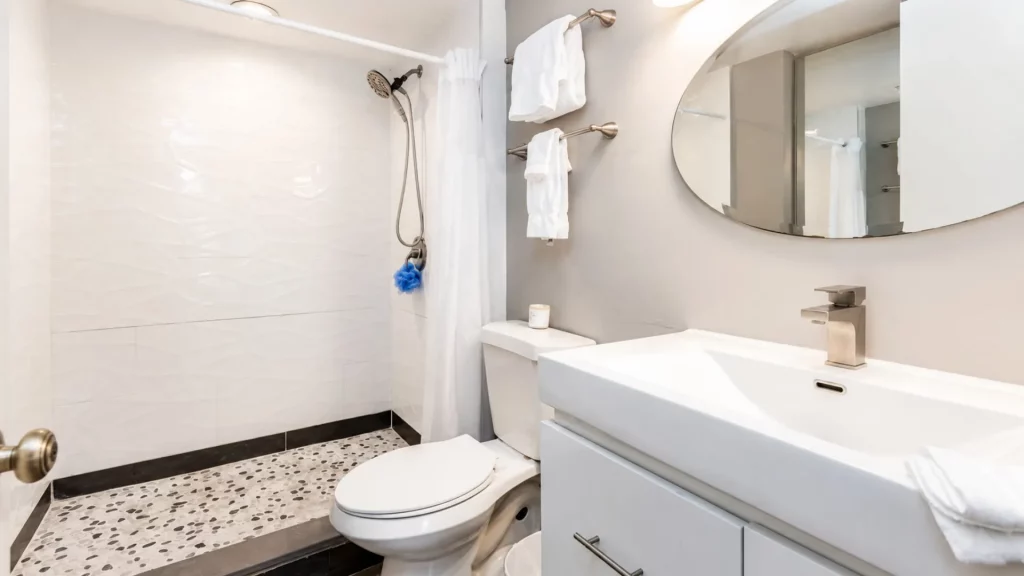 Modern bathroom with a white vanity, rectangular sink, brushed-nickel faucet, and round mirror above a toilet beside a shower with a white curtain.