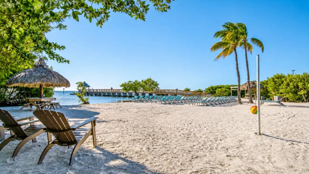 Sandy beach with wooden lounge chairs, thatched umbrella, and a pier extending into calm blue water under a clear sky.