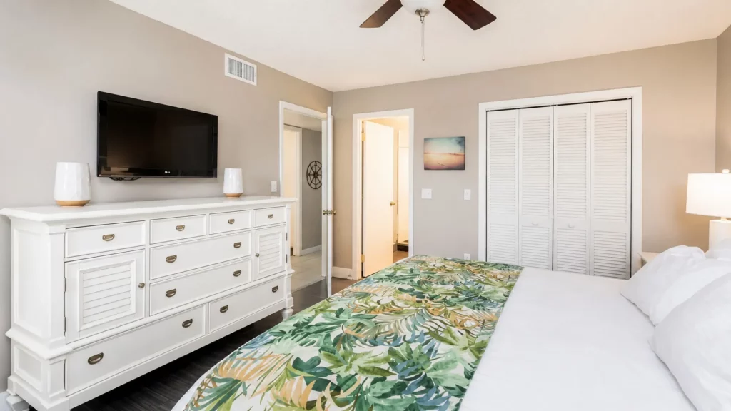 Hotel bedroom with a white dresser, wall-mounted TV, ceiling fan, and a bed with a tropical leaf runner.