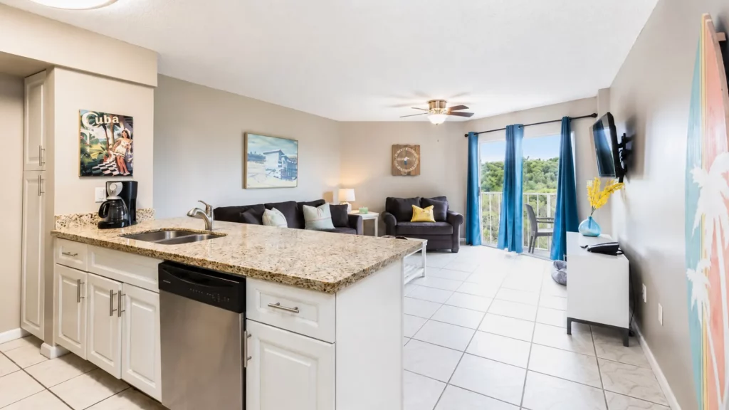 Open-plan living room and kitchen with a granite island, sink, dishwasher, coffee maker, beige cabinets, and blue-curtained sliding door to a balcony.