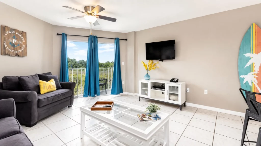 Cozy living room with a dark gray sofa, teal curtains, and a wall-mounted TV over a white media cabinet; balcony doors with a view of trees outside.