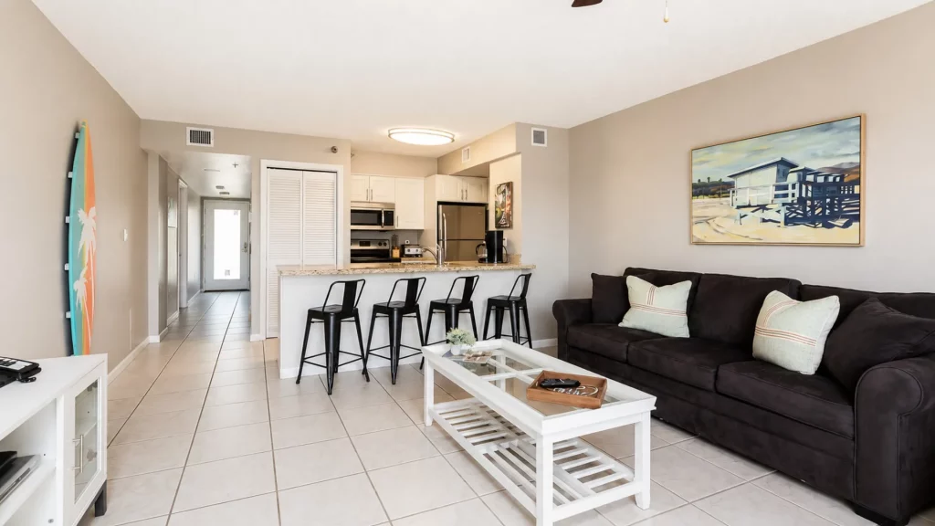 Open-concept living room and kitchen with a granite bar, five black stools, and a dark sofa with pale cushions on beige walls.