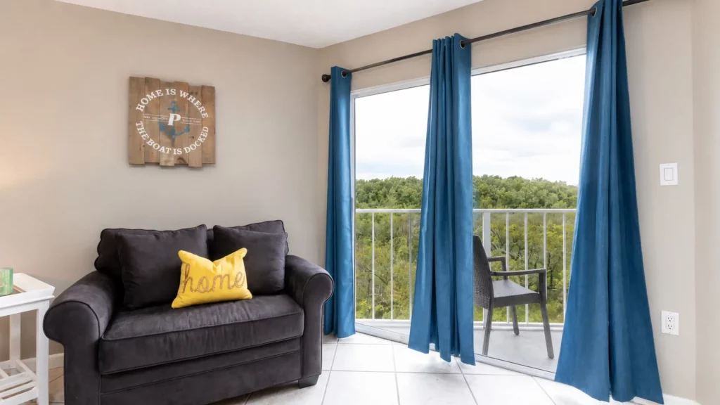 Cozy living room with a dark gray sofa, a yellow pillow, and a wooden sign on the wall reading 'Home is where the boat is docked', plus blue curtains by a balcony door.