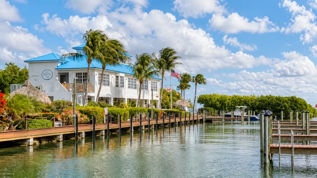 White two-story coastal house with a bright blue roof beside a marina and palm trees on the shore.