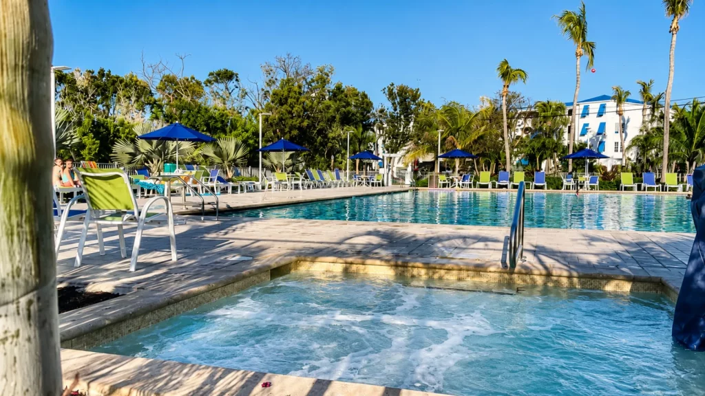 Tropical resort pool area with blue umbrellas, lime chairs, and palm trees under a bright blue sky.