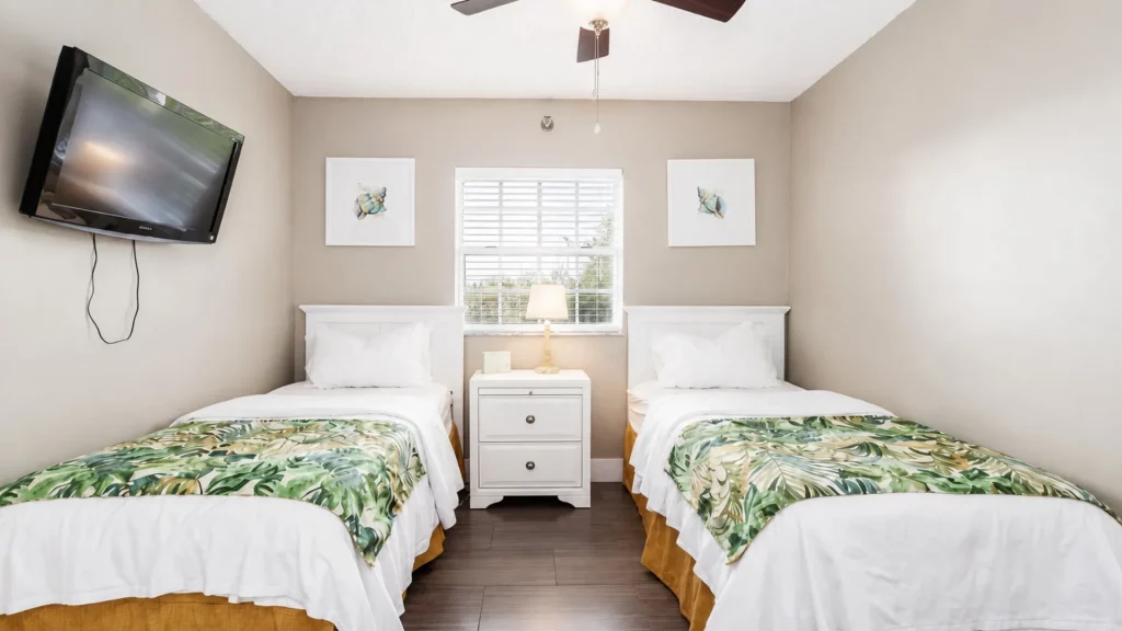 Twin bedroom with two white headboards, matching nightstand, and tropical-pattern bed runners facing a window between the beds; a wall-mounted TV on the left.