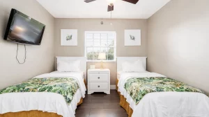 Twin bedroom with two white headboards, matching nightstand, and tropical-pattern bed runners facing a window between the beds; a wall-mounted TV on the left.