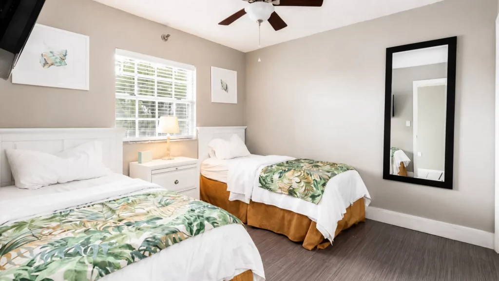 Two twin beds with white linens and tropical leaf runners, white nightstand, window with blinds, and a tall black-framed mirror on the wall.