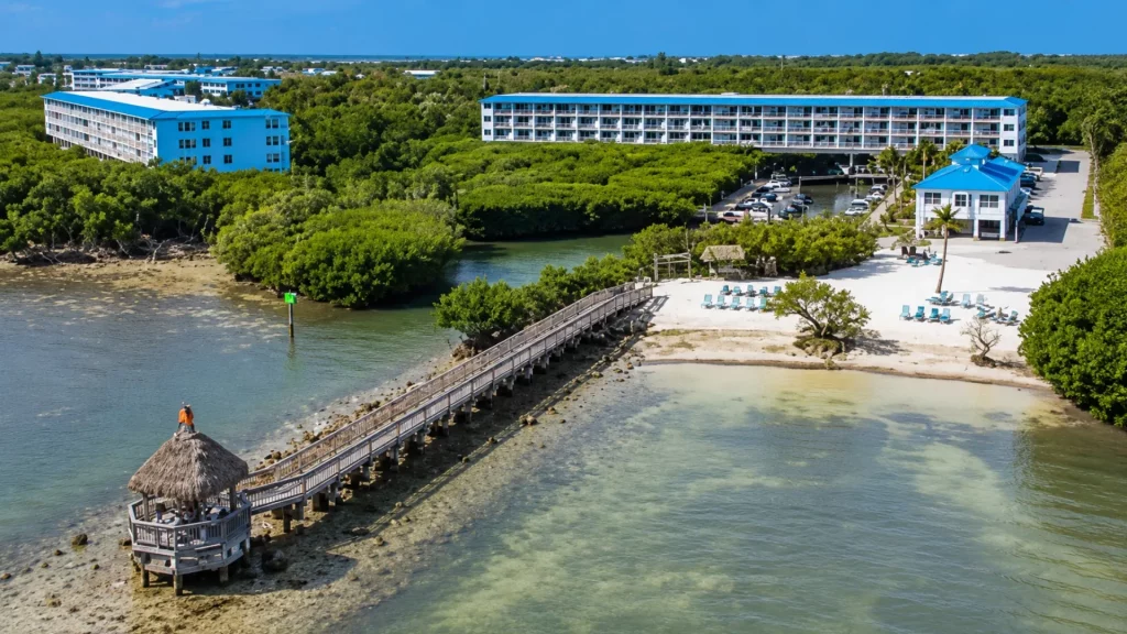 Aerial view of a coastal resort with blue buildings, a wooden boardwalk to a thatched gazebo, and a sandy beach with lounge chairs.