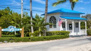 Gatehouse at Ocean Pointe with a bright blue roof, white siding, arched window, and an American flag amid tropical plants.
