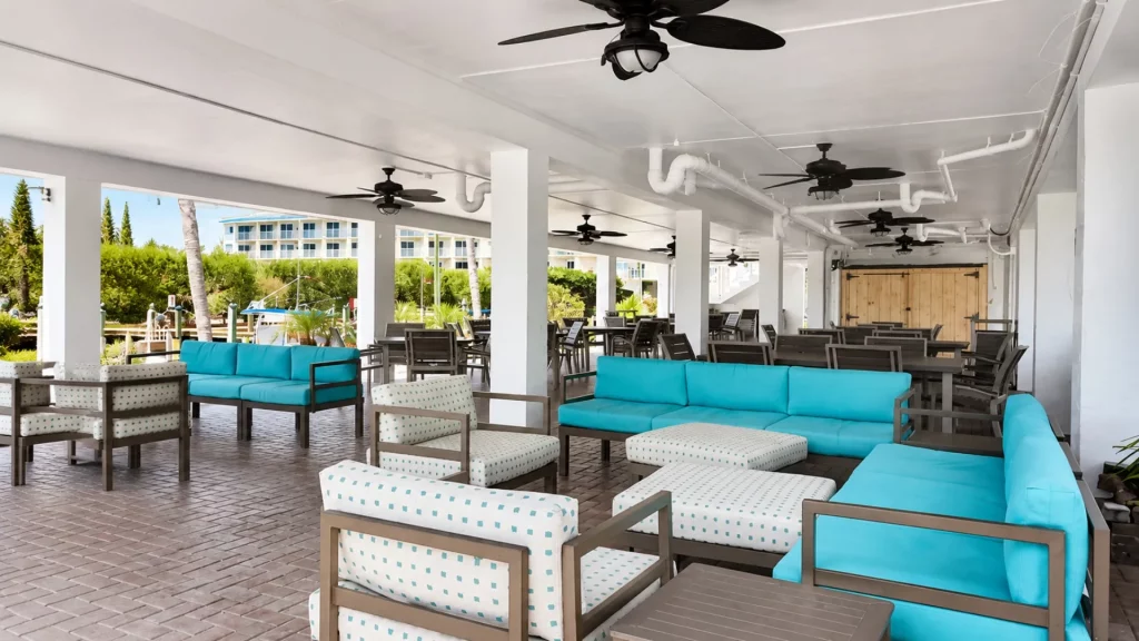 Outdoor covered lounge with turquoise-cushioned sofas and dotted chairs, wooden tables, and ceiling fans; garden and water visible beyond the columns.