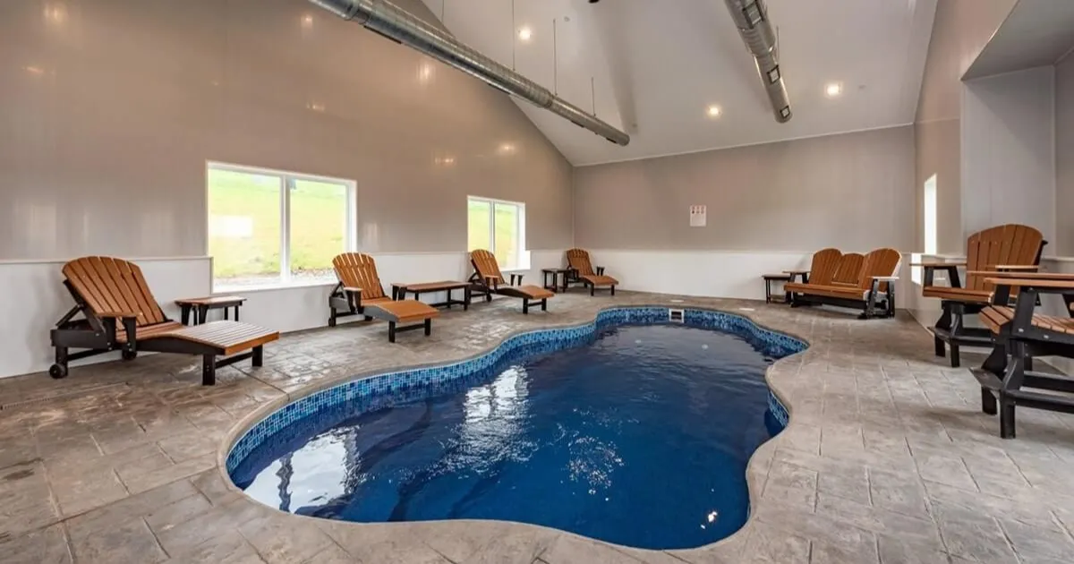 Indoor pool area with a kidney-shaped pool, blue-tiled edge, and wooden lounge chairs along the walls.
