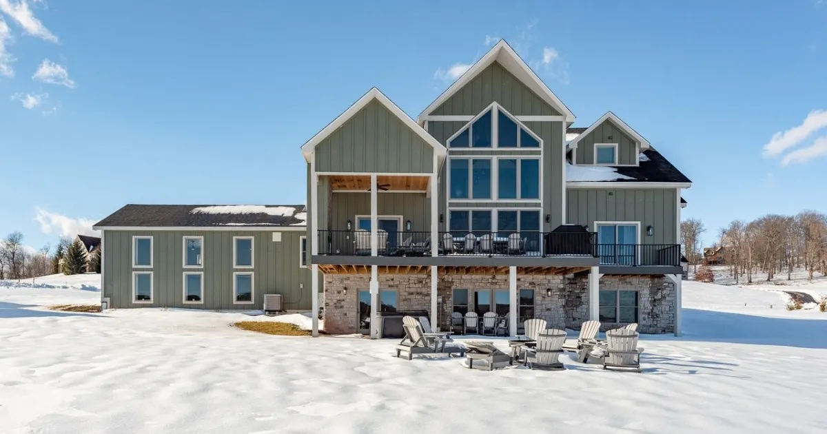 Large modern two-story house with a peaked roof, wide glass windows, and a snow-covered yard with outdoor chairs on a deck.