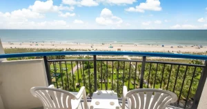 Balcony with two white plastic chairs and a small table overlooking a grassy area, sandy beach, and blue ocean under a sunny sky with scattered clouds.