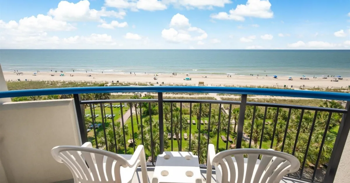 Balcony with two white plastic chairs and a small table overlooking a grassy area, sandy beach, and blue ocean under a sunny sky with scattered clouds.