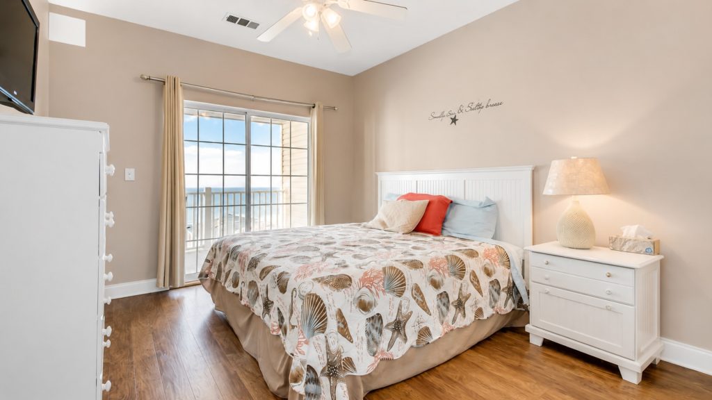 Coastal-themed bedroom with a white bed, sea-shell quilt, and a balcony door overlooking the ocean.