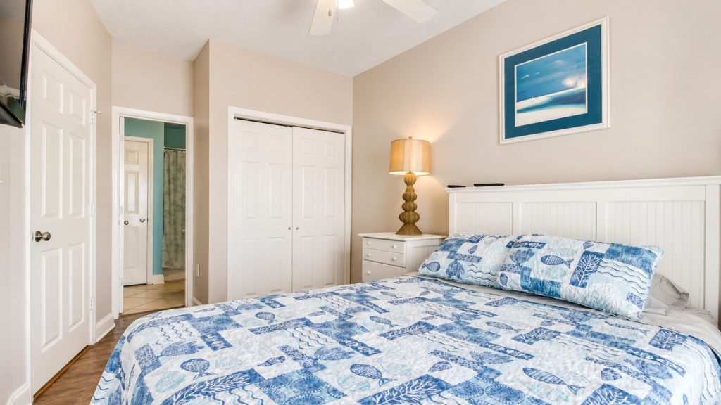 Bedroom with white headboard, blue patterned quilt, and a nightstand lamp on beige walls; framed ocean artwork above the bed.