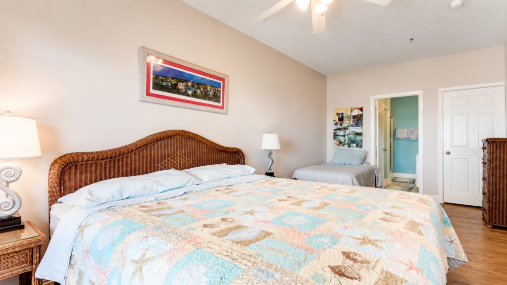 Hotel room with a large wicker headboard bed, sea-themed quilt, and a second twin bed near a bathroom doorway.