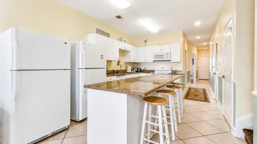 Bright white kitchen with a granite island and four wooden bar stools along the counter, beige walls in the background.
