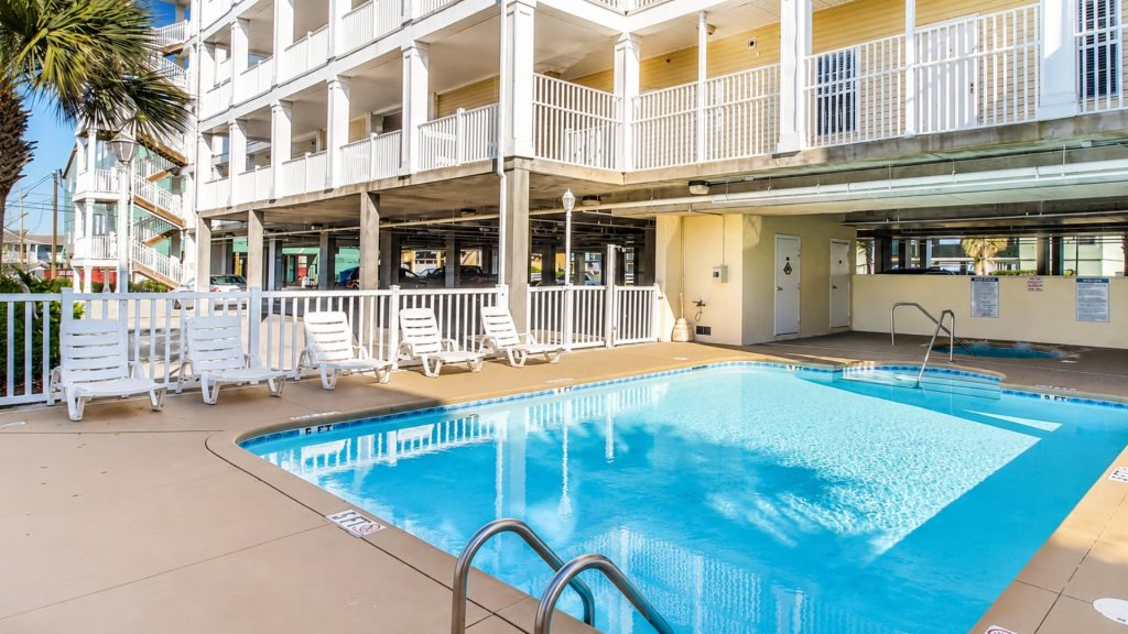 Outdoor hotel pool area with white lounge chairs along the deck and a multi-story building with balconies in the background.