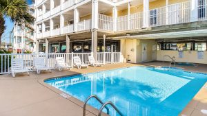 Outdoor hotel pool area with white lounge chairs along the deck and a multi-story building with balconies in the background.