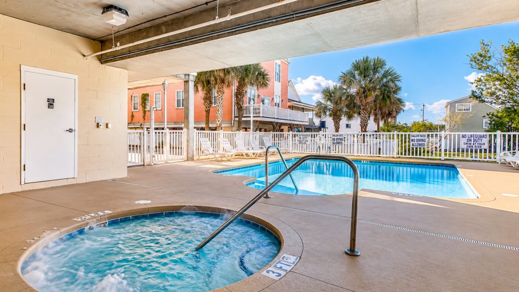 Outdoor pool area of a residential complex with a circular hot tub in the foreground, metal handrail, lounge chairs along a white fence, and palm trees under a blue sky.