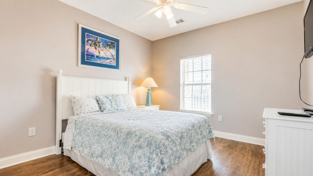 Bright bedroom with a white paneled headboard, blue floral bedding, tropical artwork, and a turquoise lamp on the nightstand beneath a ceiling fan.