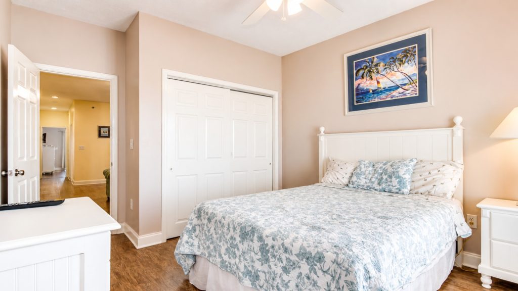 Bedroom with a white headboard, blue floral bedding, and white matching nightstands; tropical art hangs above the bed against beige walls with a hallway visible through an open door in the background.