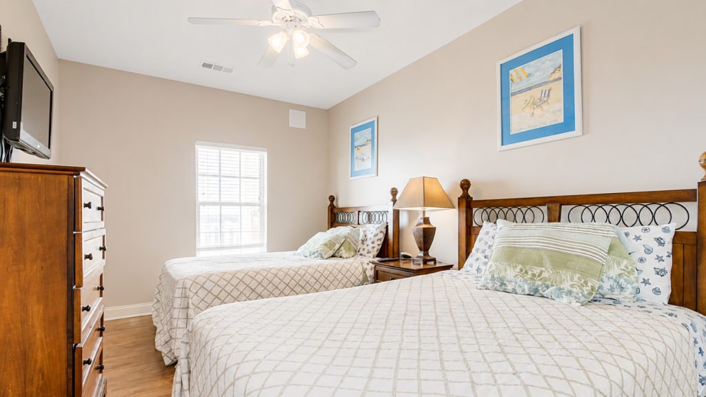 Two twin beds with wooden headboards in a bright hotel room, with a dresser, mounted TV, nightstand lamp, and beach wall art.