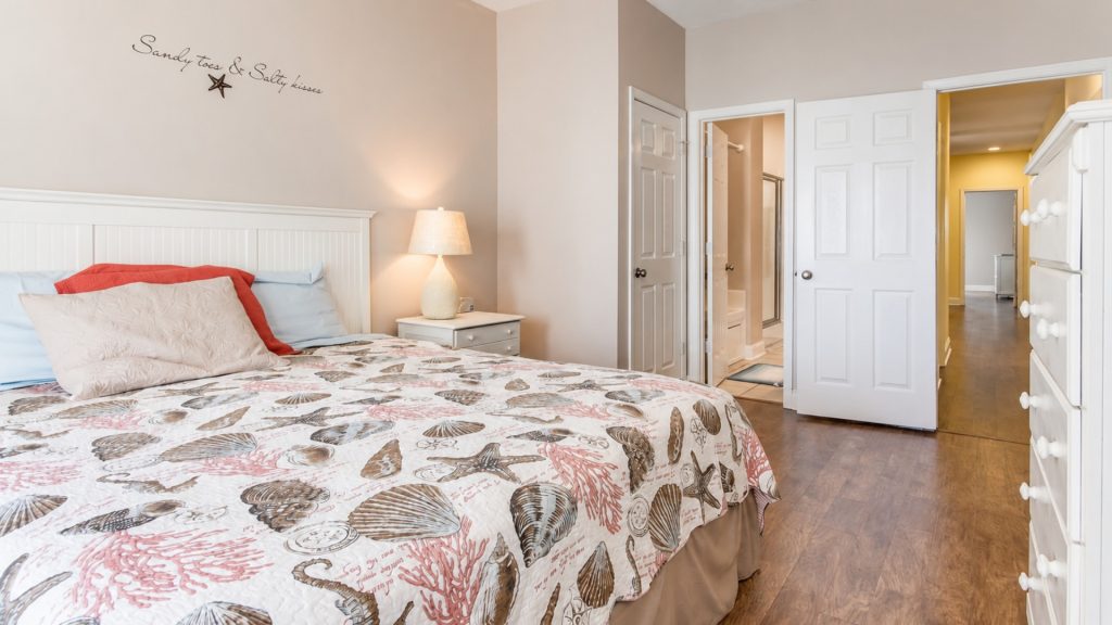 Cozy coastal bedroom with a seashell-pattern quilt, pale walls, white headboard, and a bedside lamp. Open doors lead to a bathroom and hallway.