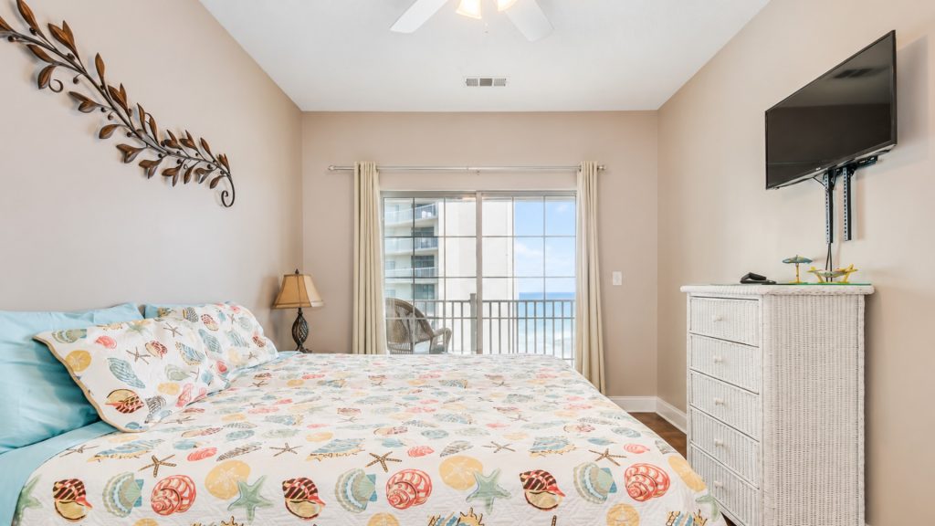 Coastal bedroom with seashell-pattern quilt and aqua pillows, right by a sliding glass balcony door showing the ocean view