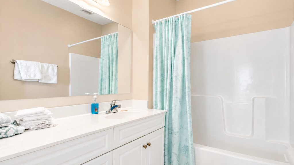 Bright bathroom with a white vanity, folded towels, blue soap dispenser, and a mint shower curtain beside a white bathtub.