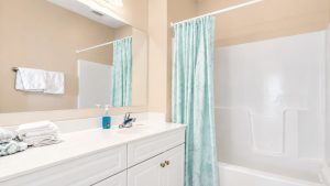 Bright bathroom with a white vanity, folded towels, blue soap dispenser, and a mint shower curtain beside a white bathtub.