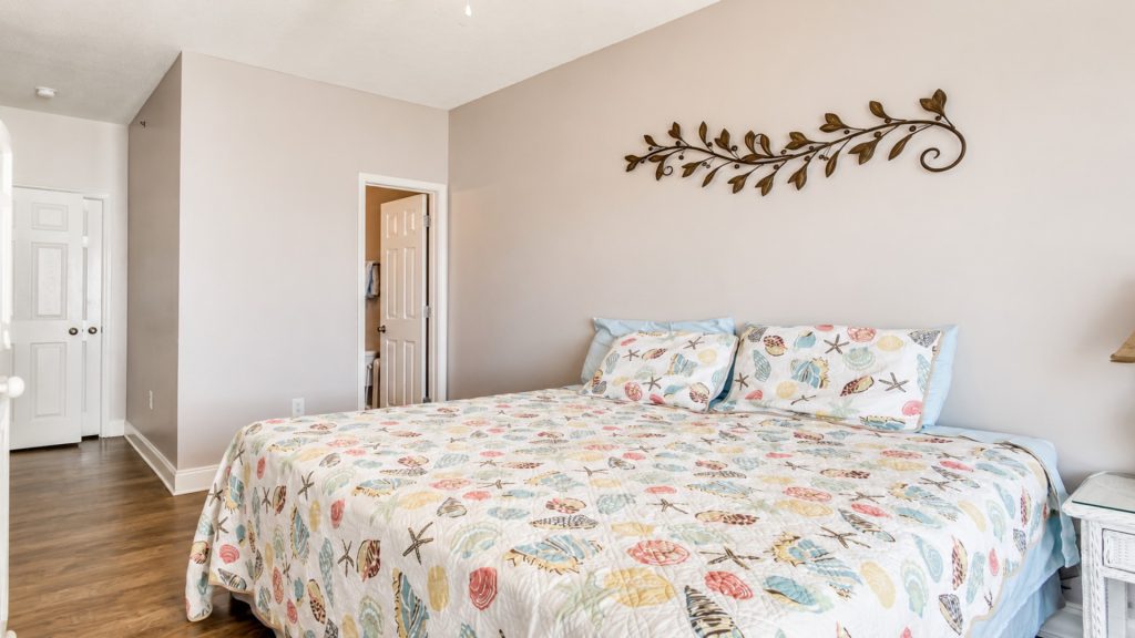 Coastal-themed bedroom with a bed dressed in a seashell and starfish quilt, two pastel pillows, beige walls, and a decorative metal vine on the wall.