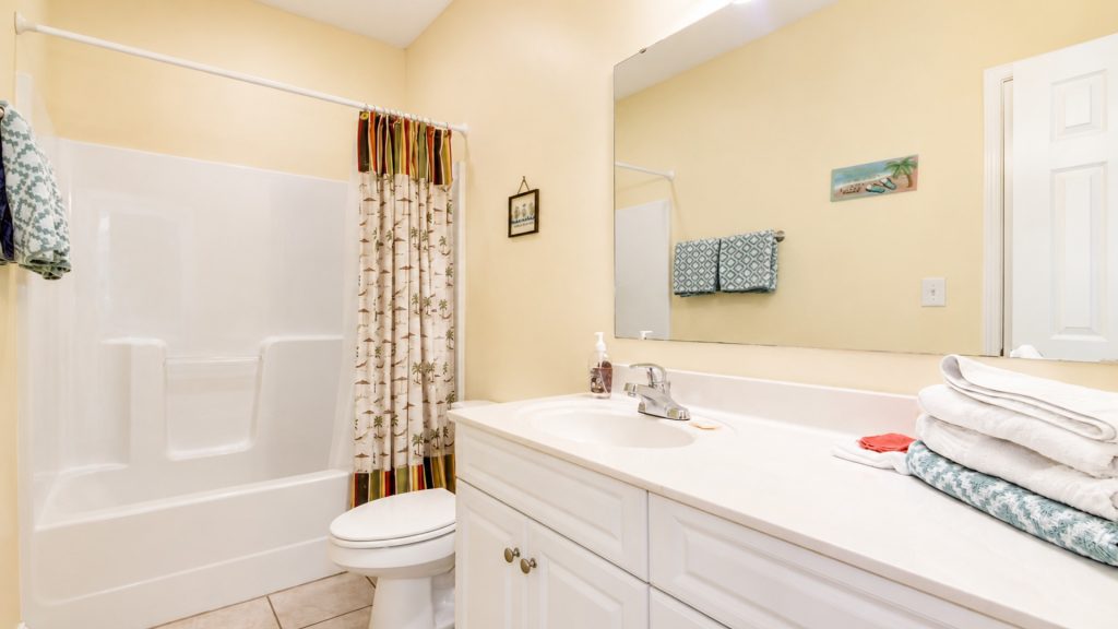 Bathroom with a white tub and shower curtain, beige walls, and a long vanity mirror above a sink with stacked towels to the right
