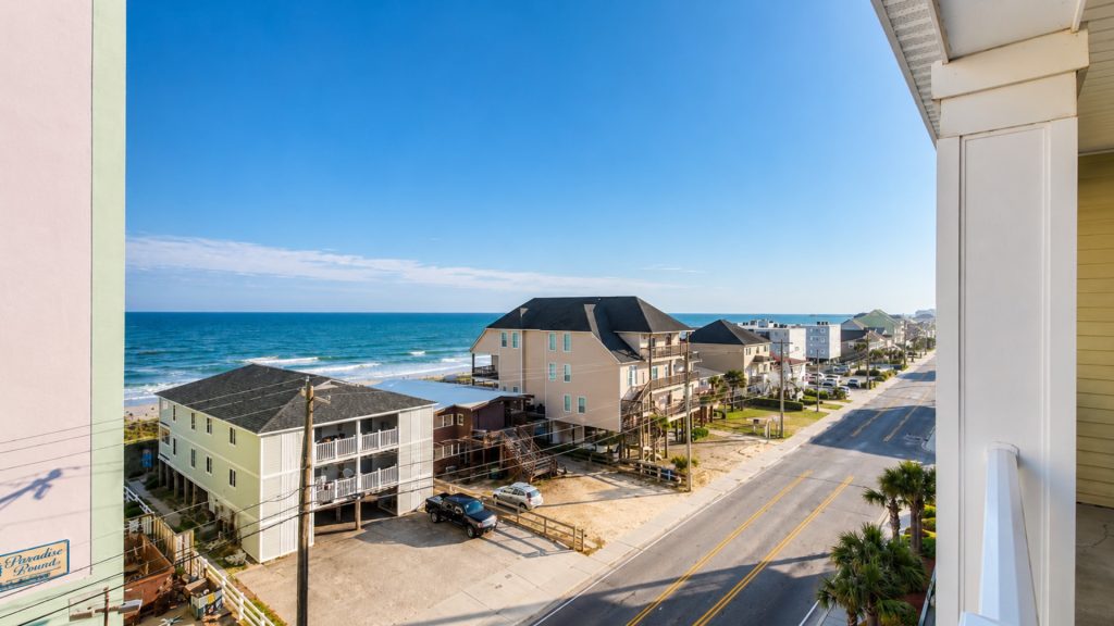 View from a balcony of a sunny coastal street with pastel beach houses and the ocean in the distance.