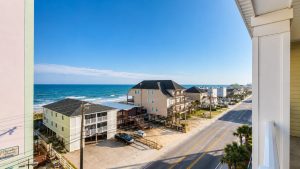 View from a balcony of a sunny coastal street with pastel beach houses and the ocean in the distance.