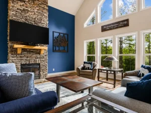 blue-bayou-Section-1 | Swack Vacations Cozy living room with a tall stacked-stone fireplace, a mounted TV, and a blue accent wall behind seating. Large windows bring in natural light.