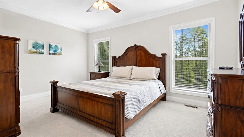 Bedroom with a large dark wood bed, matching nightstands and dresser, beige walls, and two windows with blinds.