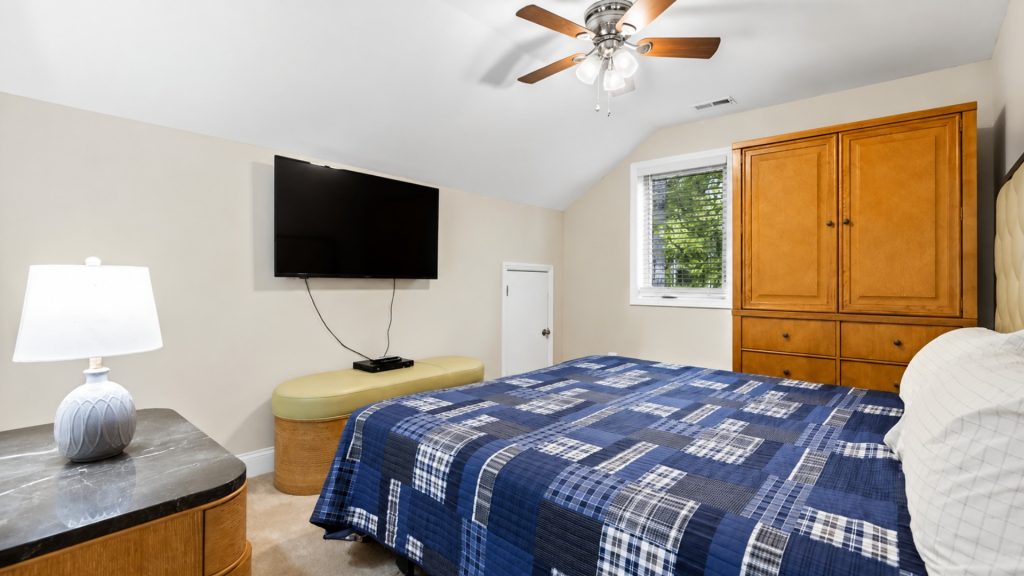 Bedroom with a wall-mounted flat-screen TV, blue plaid quilt on the bed, and a wooden cabinet against beige walls. Ceiling fan overhead; window with blinds to the right.
