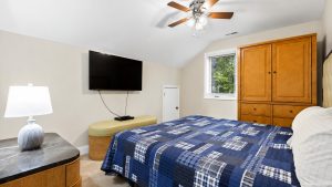 blue-bayou-bed-3-1 2 | Swack Vacations Bedroom with a wall-mounted flat-screen TV, blue plaid quilt on the bed, and a wooden cabinet against beige walls. Ceiling fan overhead; window with blinds to the right.