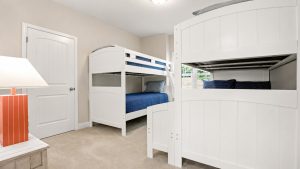 blue-bayou-bunk-room-1 | Swack Vacations Children's bedroom with white wooden bunk beds, blue bedding, a door on the left, and an orange-striped lamp on a small table.